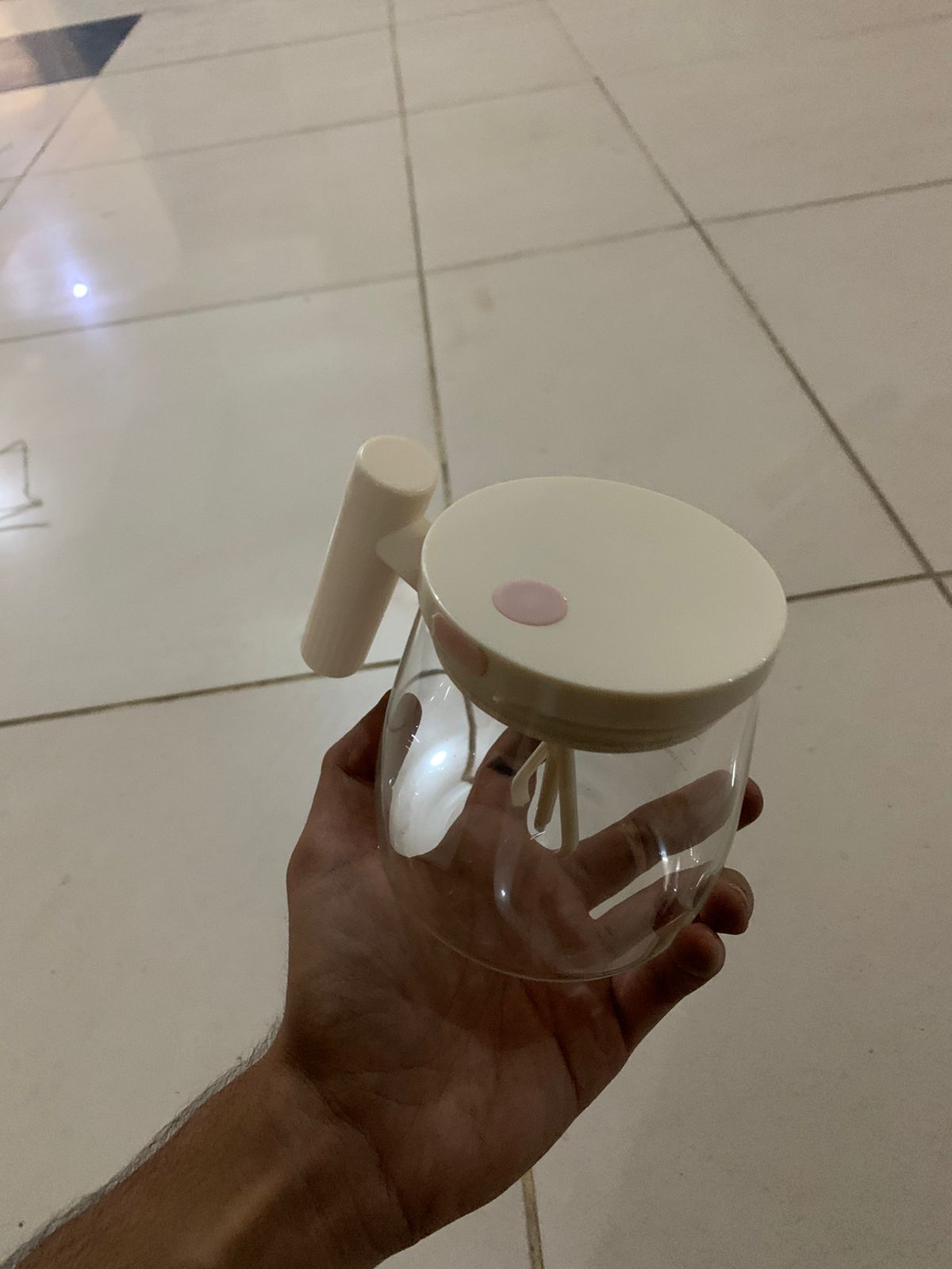 Clear glass jar with a white lid and handle held by a hand on a tiled floor.
